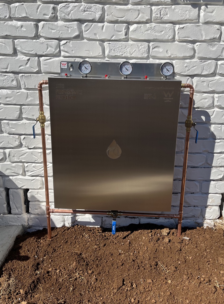 Stainless whole-house water filter with copper pipework on a painted brick wall